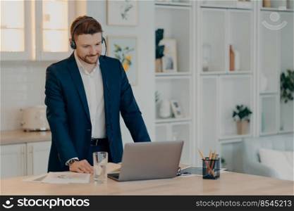 Smiling business professional wearing suit and headset, listening attentively to his colleague during video call on laptop, standing near table in kitchen while taking part in online meeting. Focused young businessman standing near dining table and working on laptop