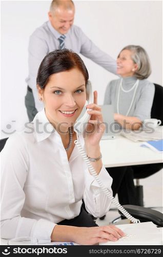 Smiling business secretary calling phone happy colleagues in background