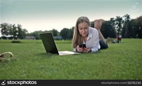 Smiling business woman lying on green grass barefoot on park&acute;s lawn and typing on the cellphone. Executtive working outddoor