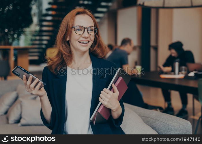 Smiling business woman with modern smartphone in hand, sitting on couch in lounge zone, holding laptop with notebook, smiling and looking around while waiting for meeting with client in office space. Smiling woman holding laptop, notebook and modern smartphone while waiting for business partner