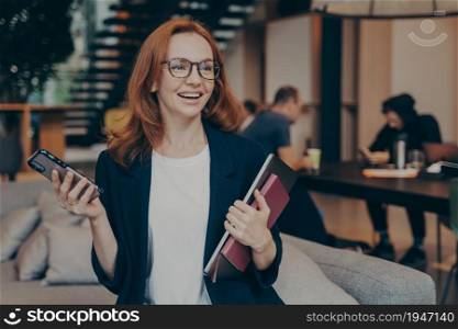 Smiling business woman with modern smartphone in hand, sitting on couch in lounge zone, holding laptop with notebook, smiling and looking around while waiting for meeting with client in office space. Smiling woman holding laptop, notebook and modern smartphone while waiting for business partner