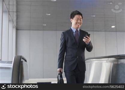 Smiling businessman coming up the escalator and looking down at his phone