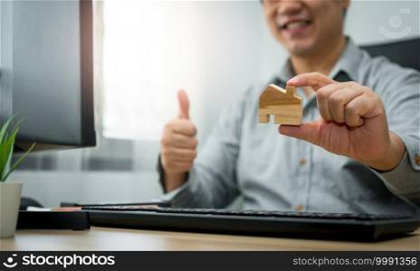 Smiling businessman holding a wooden house in home workspace. The concept of working from home until the end of Coronavirus  COVID-19  quarantine, happily freelance working lifestyle