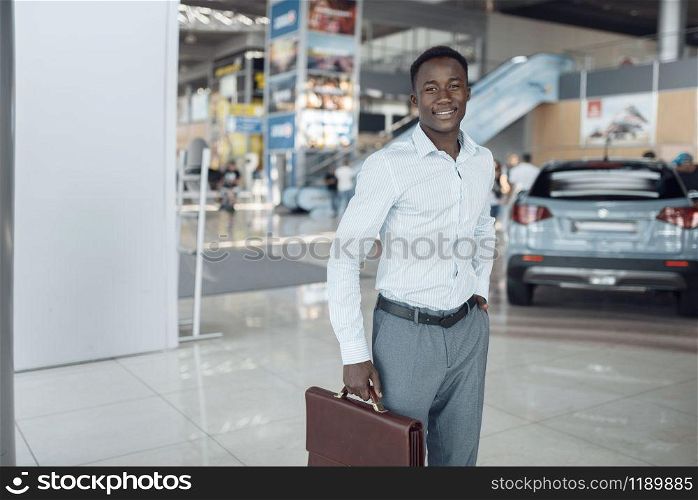 Smiling businessman holds briefcase in car showroom. Successful business person on motor show, black man in formal wear. Smiling businessman holds briefcase, car showroom