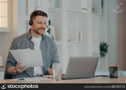 Smiling businessman in casual wear and headset sitting and looking at laptop screen while working at home, making notes and analyzing documents during online meeting. Remote job concept. Happy young guy in casual clothes during meeting online