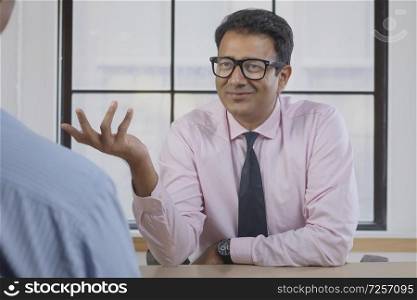 Smiling businessman in eyeglasses discussing work with client sitting at his desk in office