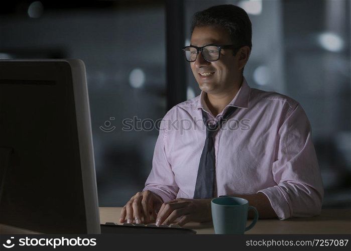 Smiling businessman in eyeglasses working on computer sitting late in office with a cup of coffee on table