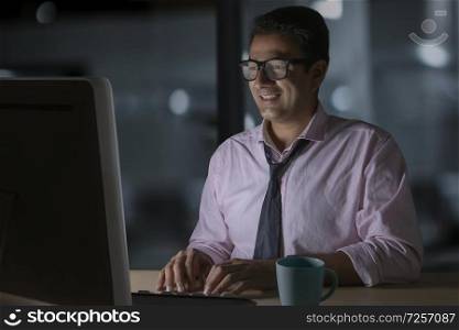 Smiling businessman in eyeglasses working on computer sitting late in office with a cup of coffee on table