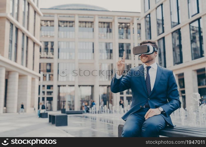 Smiling businessman sitting on bench in town square with VR glasses on, viewing information in virtual reality, pointing upwards with his finger, testing new innovative method for business. Smiling businessman with stubble sitting on bench in town square with VR glasses on