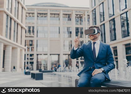 Smiling businessman sitting on bench in town square with VR glasses on, viewing information in virtual reality, pointing upwards with his finger, testing new innovative method for business. Smiling businessman with stubble sitting on bench in town square with VR glasses on