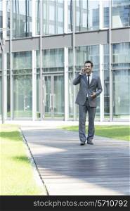 Smiling businessman using mobile phone while walking on path outside office