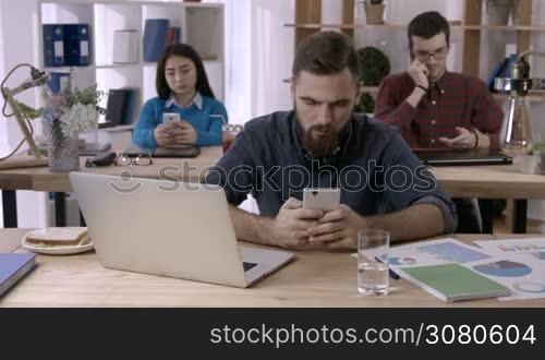 Smiling businessman with beard browsing social networks on mobile phone in open space office as multi-ethnic colleagues busy with smartphones on background. Young freelancer texting on cellphone.