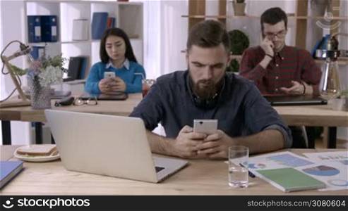 Smiling businessman with beard browsing social networks on mobile phone in open space office as multi-ethnic colleagues busy with smartphones on background. Young freelancer texting on cellphone.