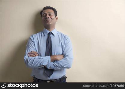 Smiling businessman with eyes closed standing with his back against a wall with arms crossed