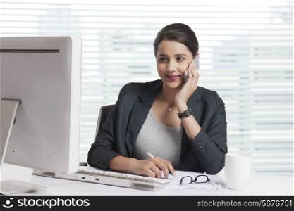 Smiling businesswoman answering smart phone while looking at computer in office
