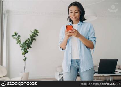 Smiling businesswoman holding smartphone typing message in corporate app standing in modern office space. Happy female employee using phone planning work time, browsing working news at workplace.. Smiling businesswoman holding smartphone types message in corporate app reads working news in office