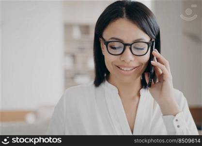 Smiling businesswoman in glasses talks on phone. Happy young female businessperson answering call, talking on smartphone with client in office or enjoying corporate mobile conversation.. Smiling businesswoman wearing glasses talking on smartphone with client, answering call in office
