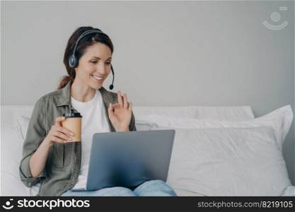 Smiling businesswoman in headset look at laptop screen talk on video call with client or customer showing okay gesture. Female employee in headphones conducts online consultation from home. Remote job. Businesswoman in headset talk by video call on laptop with client showing okay gesture. Remote job