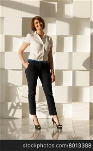 Smiling businesswoman in high heel shoes looking at camera while holding hand in pocket. Modern white wall on background. Smiling woman looking at camera while holding hand in pocket