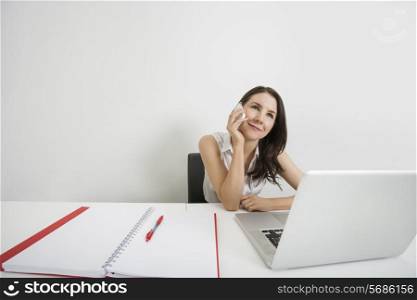 Smiling businesswoman looking away while answering cell phone at desk in office
