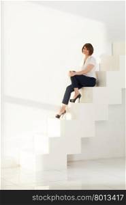 Smiling businesswoman sitting on steps of modern ladder. Elegant adult woman smiling while sitting on steps