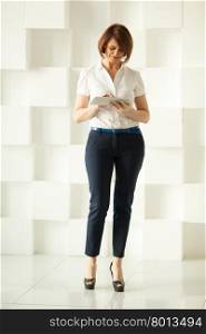 Smiling businesswoman standing against white wall while looking at tablet in her hand. Businesswoman standing against white wall while looking at tablet