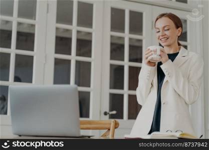 Smiling businesswoman standing at office desk enjoying pause break with cup of morning coffee during work at laptop. Happy young female employee drinking tea from mug in the workplace.. Smiling female employee standing at office desk enjoying break with coffee cup during work at laptop