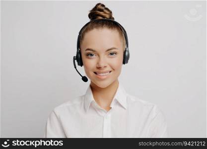 Smiling call center worker in white shirt, hair in bun, black headset, eager to serve and assist customers, stands alone against grey background.