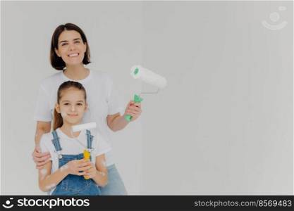 Smiling caring mother embraces daugher, stands with paint rollers, paint walls in house, girl has dirty face, dressed in denim overalls, poses in empty room, copy space aside. Home repair concept