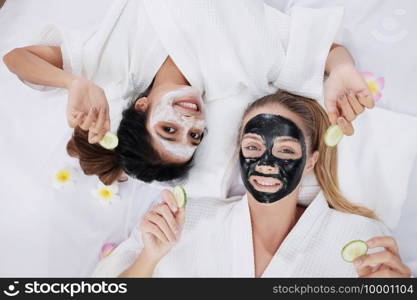 Smiling Caucasian and Asian two girls friends is holding Sliced   cucumber and relaxing with facial masks for skine care on white bed together. Beauty salon and lifestyle of lesbian Concept.