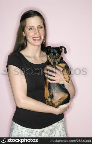 Smiling Caucasian woman holding Miniature Pinscher dog standing against pink background.