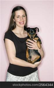 Smiling Caucasian woman holding Miniature Pinscher dog standing against pink background.