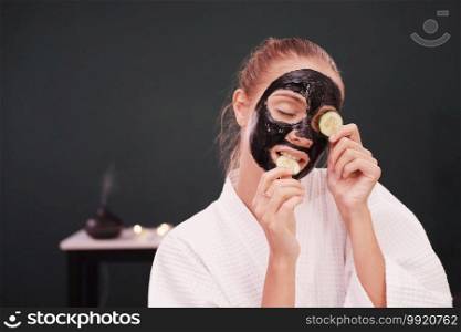 Smiling Caucasian woman in white bathrobe is holding cucumber slices and black facial mask on face for treatment skine care at spa.