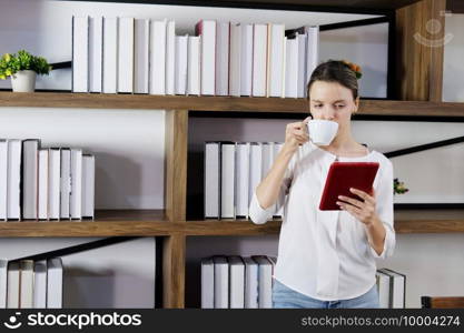 Smiling Caucasian woman is drinking coffee in the library and reading online news on tablet for knowledge and work with internet at university or modern office. Technology and education concept