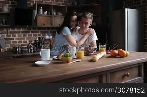 Smiling charming woman embracing handsome hipster man and using smart phone together in the kitchen during breakfast in the morning. Slow motion. Steadicam stabilized shot.