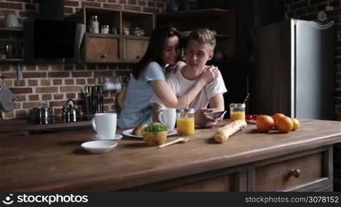 Smiling charming woman embracing handsome hipster man and using smart phone together in the kitchen during breakfast in the morning. Slow motion. Steadicam stabilized shot.
