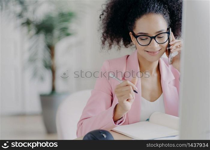 Smiling charming woman with curly hair, writes down information in notebook, gets consultancy via cellphone, wears glasses and elegant suit, busy working, owns business company, solves problems
