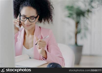 Smiling charming woman with curly hair, writes down information in notebook, gets consultancy via cellphone, wears glasses and elegant suit, busy working, owns business company, solves problems