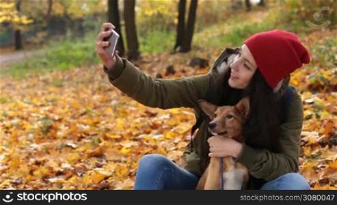 Smiling charming woman with her dog taking selfie on smartphone in colorfur autumn park. Cheerful hipster girl in stylish outfit and her cute puppy sitting on yellow fallen leaves and making self portrait photography in indian summer.