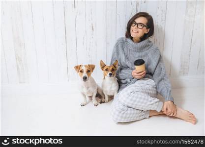 Smiling cheerful young female wears warm woolen sweater, square eyewear, drinks hot beverage, relaxes on floor and her two favourite pets, look directly into camera. People and animals conecpt