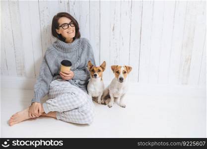 Smiling cheerful young female wears warm woolen sweater, square eyewear, drinks hot beverage, relaxes on floor and her two favourite pets, look directly into camera. People and animals conecpt