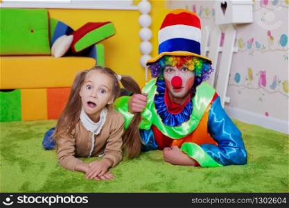 Smiling child and the clown lie on a carpet in the nursery. Colourful sofa, nesting box and fence on the background.