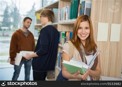 Smiling college student holding books with classmates standing in library