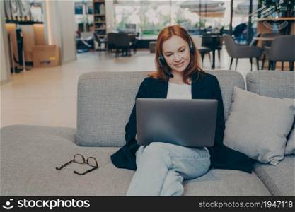 Smiling confident redhead lady employer, having business webinar online, participating in financial convection and entrepreneurship, using laptop and wireless headset, sitting on sofa in coffee house. Smiling confident redhead lady using laptop and wireless headset, sitting on sofa in coffee house