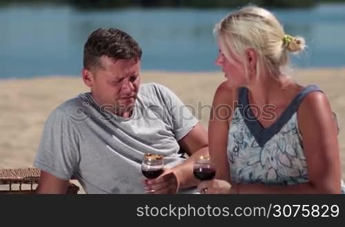Smiling couple drinking red wine from crystal glasses and chatting during picnic on the beach