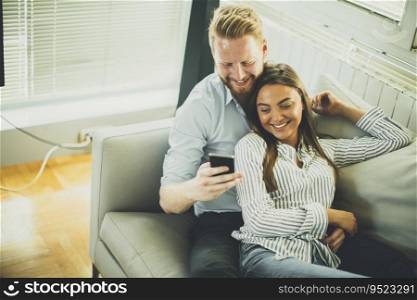 Smiling couple in casual outfit, sitting on the couch at the living room while watching something at mobile phone.