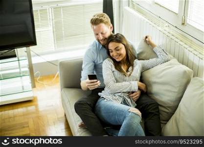 Smiling couple in casual outfit, sitting on the couch at the living room while watching something at mobile phone.