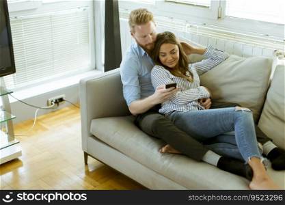 Smiling couple in casual outfit, sitting on the couch at the living room while watching something on mobile phone
