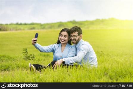 Smiling couple in love sitting on the grass taking selfies, Young couple in love taking a selfie in the field, People in love taking selfies in the field with their smartphone