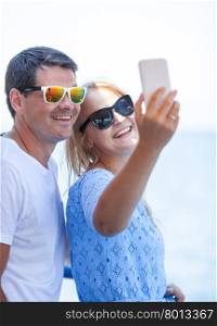 Smiling couple in sunglasses making happy summer selfie with cell against blue sea and sky background. Cheerful couple in sunglasses taking mobile selfie